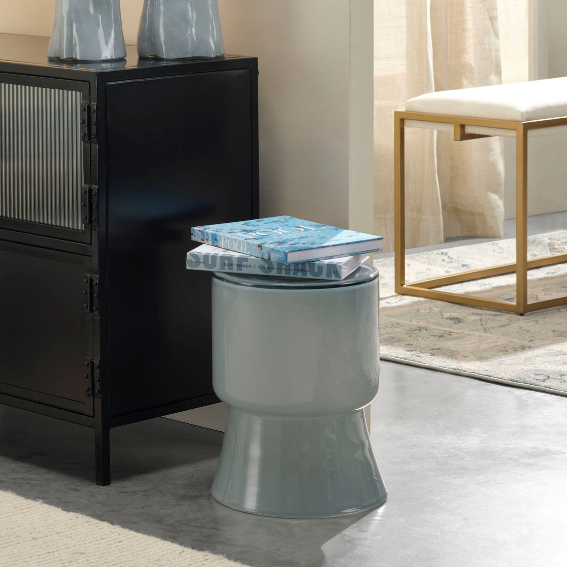 Gray stool with a book on top in a room setting with furniture and decor.
